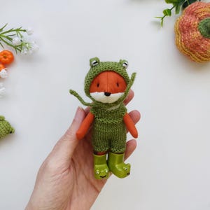 Puede incluir: Una muñeca de zorro hecha a mano con un disfraz de rana verde, que incluye una capucha y botas. El zorro tiene brazos naranjas y una cara blanca. También hay una pequeña muñeca de rana de ganchillo verde. El fondo incluye una calabaza tejida y detalles florales.