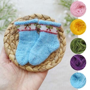 May include: A pair of light blue knitted baby socks with a white and red cherry blossom design on the cuffs. The socks are displayed on a small woven mat, with colorful yarn samples in pink, yellow, green, purple, and blue in the background.