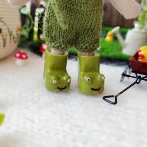Könnte beinhalten: Eine Puppe mit grünen Froschstiefeln und einer grünen Strickhose. Ein kleiner roter Wagen mit einer gelben Quietscheente befindet sich hinter der Puppe. Miniatur-Dekorationen mit roten und weißen Pilzen sind verstreut.
