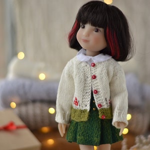 May include: A doll wearing a white knitted cardigan with red mushroom embroidery and a green skirt. The doll has dark hair with red highlights and is looking to the side.