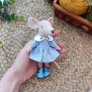 May include: A small, handmade mouse doll wearing a light blue dress with a white collar and yellow accents. The dress has a bee design on the collar. The mouse is holding a small, white stick in its hand.