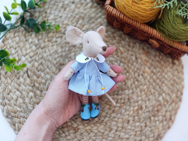 May include: A small, handmade mouse doll wearing a light blue dress with a white collar and yellow accents. The dress has a bee design on the collar. The mouse is holding a small, white stick in its hand.