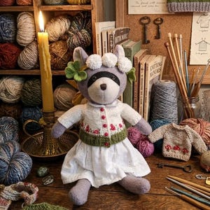 May include: A plush raccoon doll wearing a white knit cardigan with red floral embroidery, a green ribbed hem, and a white eyelet skirt. The raccoon has a white floral and green leaf crown on its head. The doll is positioned among yarn balls, knitting needles, and miniature sweaters.