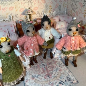 May include: Four small, gray, knitted mouse figurines wearing different colored sweaters and hats. The mice are standing on a patterned rug in a dollhouse bedroom setting.