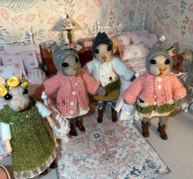 May include: Four small, gray, knitted mouse figurines wearing different colored sweaters and hats. The mice are standing on a patterned rug in a dollhouse bedroom setting.