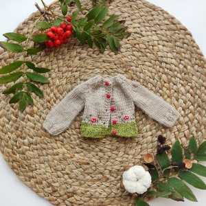 Op de afbeelding: Een kleine, handgebreide beige cardigan met lange mouwen en rode knopen. De onderste zoom is groen, met rode paddenstoelaccenten. De cardigan wordt gepresenteerd op een geweven, cirkelvormige mat, met takjes bladeren en bessen.