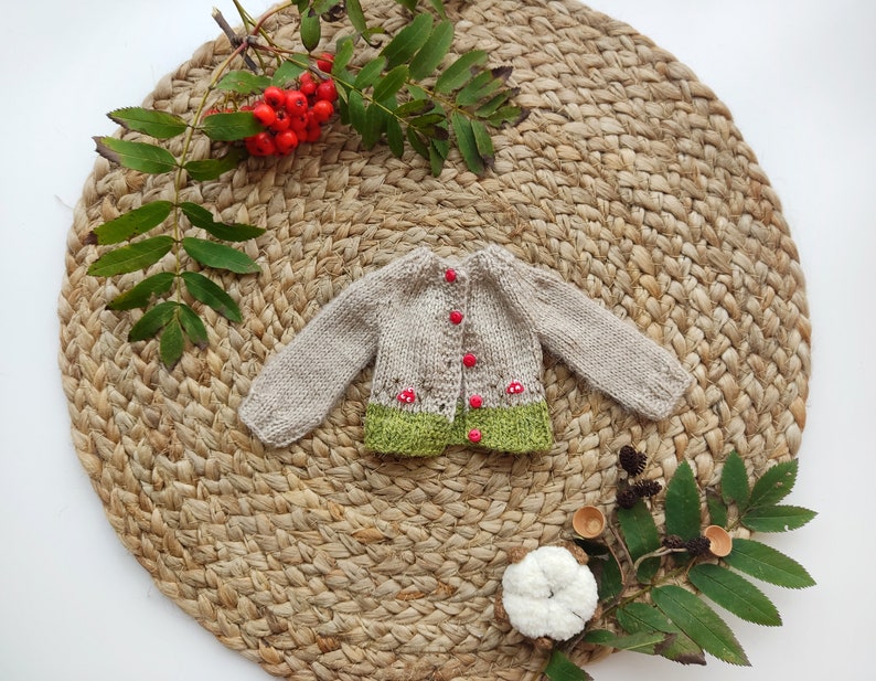 Op de afbeelding: Een kleine, handgebreide beige cardigan met lange mouwen en rode knopen. De onderste zoom is groen, met rode paddenstoelaccenten. De cardigan wordt gepresenteerd op een geweven, cirkelvormige mat, met takjes bladeren en bessen.