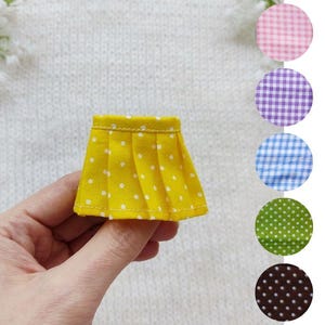 May include: A miniature yellow pleated skirt with white polka dots is held in a hand. The skirt is displayed next to fabric swatches in pink, purple, blue, green, and brown gingham and polka dot patterns.