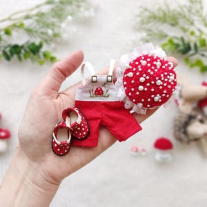 Pode incluir: É apresentado um conjunto vermelho em miniatura com bolinhas brancas, incluindo macacão e sapatos. Também é apresentado um acessório em forma de cogumelo vermelho com bolinhas brancas e uma borda de renda branca. Os itens são provavelmente para uma boneca ou um projeto de artesanato semelhante.