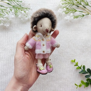 May include: A handmade doll resembling a hedgehog, with a plush brown fur head and a soft fabric body. The doll wears a pink and white sweater, a matching skirt, and pink shoes with floral accents. The doll is approximately 4 inches tall.