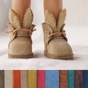 Könnte beinhalten: Ein Paar hellbraune Wildlederstiefel mit Hasenohren-Applikationen. Die Stiefel haben Schnürsenkel und sind am Knöchel gebunden.