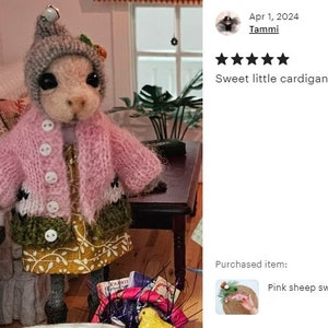 May include: A pink knitted cardigan with white and green accents for a small doll. The cardigan has a button closure and is worn over a yellow and white floral dress.