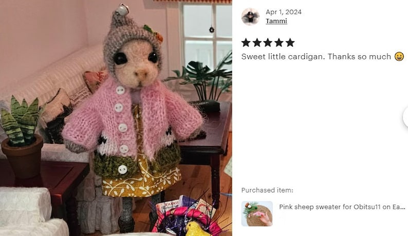 May include: A pink knitted cardigan with white and green accents for a small doll. The cardigan has a button closure and is worn over a yellow and white floral dress.