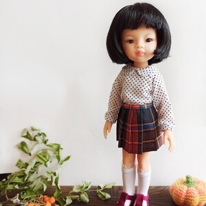 May include: A doll with black hair and a white shirt with black polka dots. The doll is wearing a red, black, and blue plaid skirt and white socks. The doll is wearing burgundy shoes with a strap. The doll is standing on a wooden surface with a small orange pumpkin next to it.