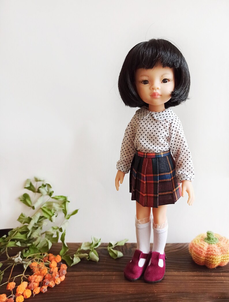 May include: A doll with black hair and a white shirt with black polka dots. The doll is wearing a red, black, and blue plaid skirt and white socks. The doll is wearing burgundy shoes with a strap. The doll is standing on a wooden surface with a small orange pumpkin next to it.