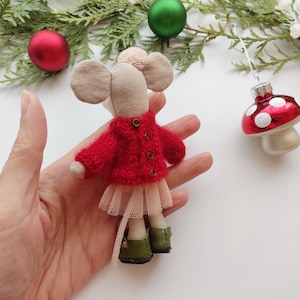 Op de afbeelding: Een kleine, handgemaakte muispop met een rode gebreide trui, een tule rok en groene schoenen. De pop wordt in een hand gehouden tegen een witte achtergrond.