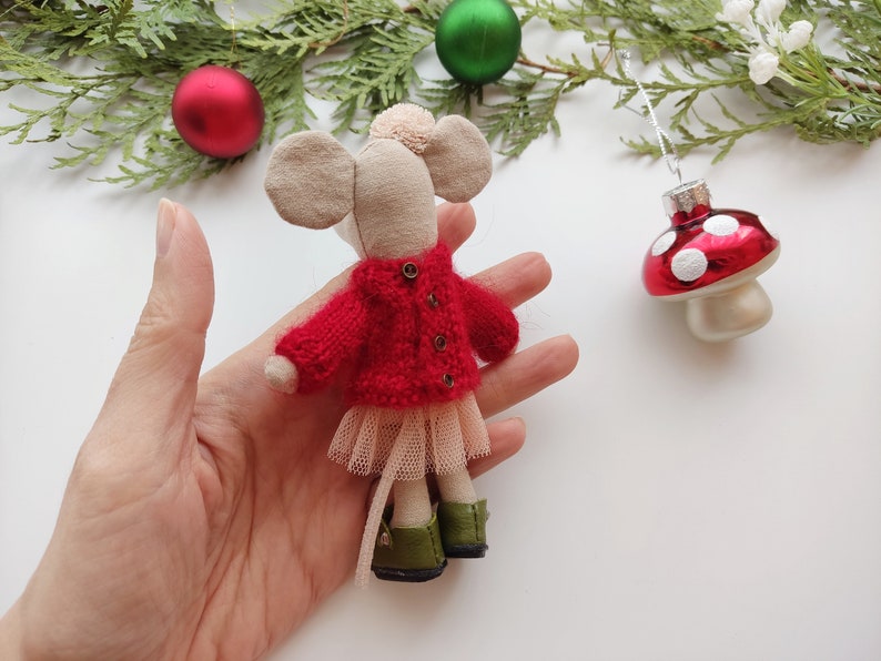 Op de afbeelding: Een kleine, handgemaakte muispop met een rode gebreide trui, een tule rok en groene schoenen. De pop wordt in een hand gehouden tegen een witte achtergrond.