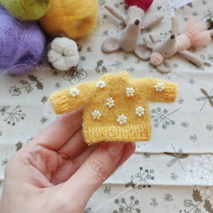 May include: A yellow knitted sweater with white daisy embellishments. The sweater is small enough to fit in the palm of a hand.