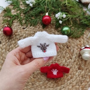 May include: A white knitted sweater with a brown reindeer design and a red pom-pom nose. The sweater is small and is being held in a hand. There is a red and white knitted sweater in the background.