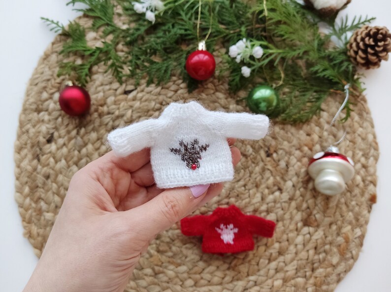 May include: A white knitted sweater with a brown reindeer design and a red pom-pom nose. The sweater is small and is being held in a hand. There is a red and white knitted sweater in the background.