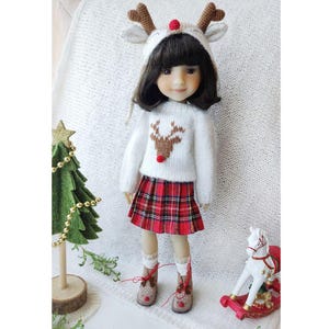 Może przedstawiać: Lalka ubrana w świąteczny strój bożonarodzeniowy. Lalka ma na sobie biały sweter z motywem renifera, czerwoną spódnicę w kratę i czapkę z rogami renifera. Mała choinka i konik na biegunach znajdują się w tle.
