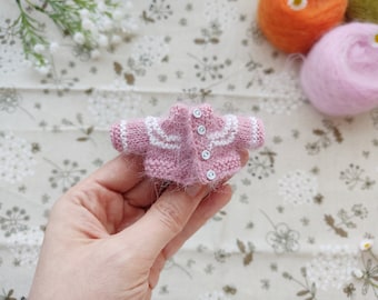 May include: A tiny pink knitted cardigan with white trim and button closures.