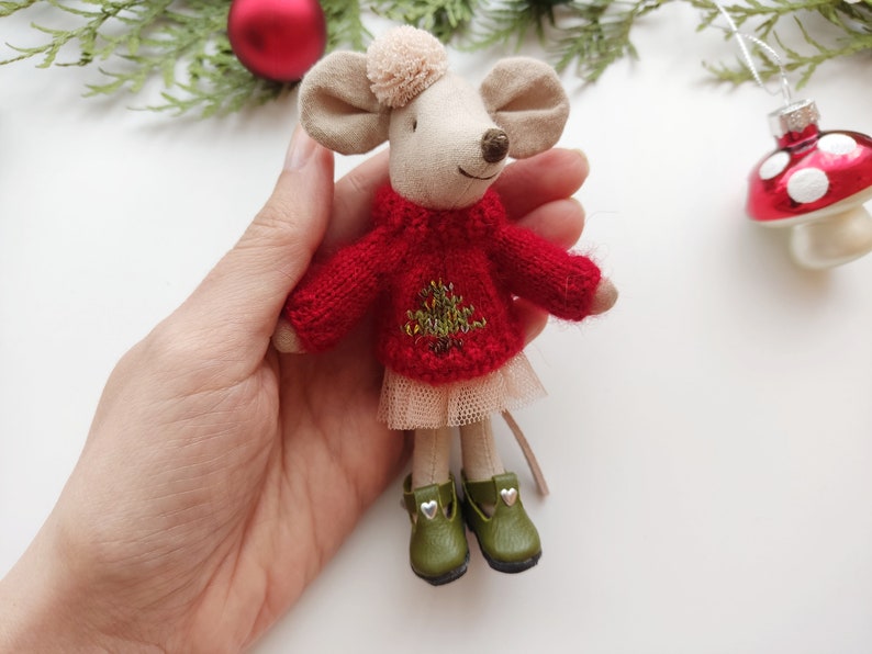 Op de afbeelding: Een kleine, handgemaakte muispop die een rode gebreide trui draagt met een groen geborduurd kerstboommotief, een beige tule rok en groene schoenen met zilveren hartjes.