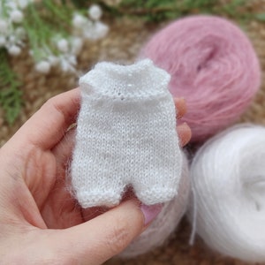 May include: A white knitted baby onesie with a delicate stitch pattern. The onesie is held in a hand, with yarn balls in the background.