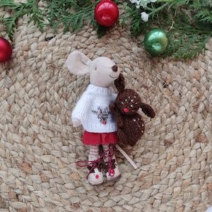 May include: A white knitted sweater with a red reindeer design, worn by a plush mouse doll holding a brown plush reindeer. The doll is standing on a woven brown mat with red and green ornaments.