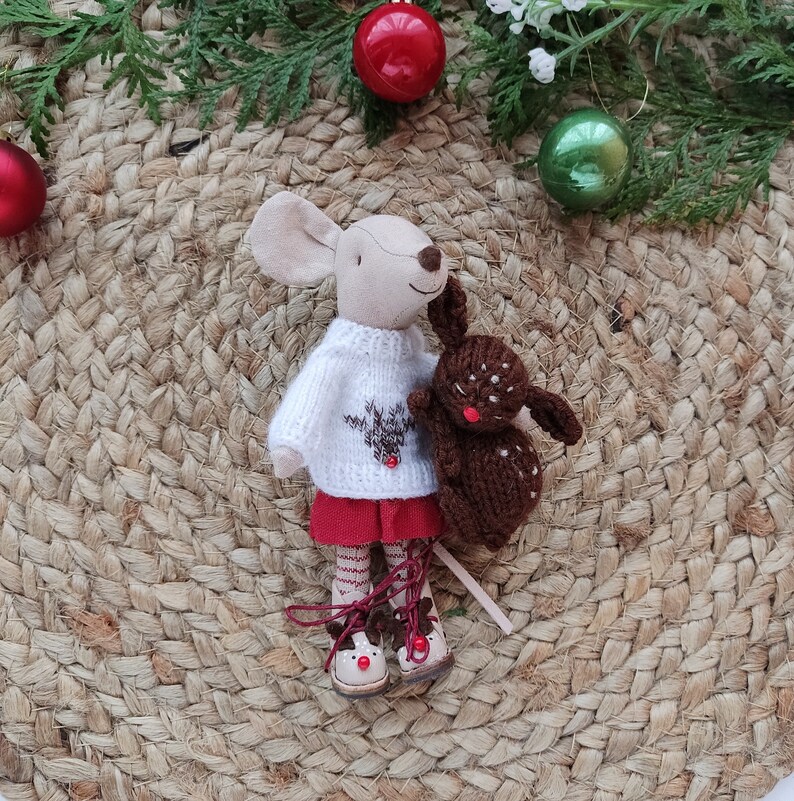 May include: A white knitted sweater with a red reindeer design, worn by a plush mouse doll holding a brown plush reindeer. The doll is standing on a woven brown mat with red and green ornaments.