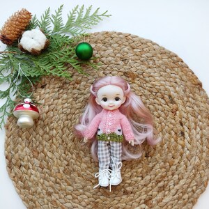 May include: A pink and green knitted sweater with a white collar and a green leaf design. The sweater is worn by a doll with pink hair and blue eyes. The doll is sitting on a brown woven mat.