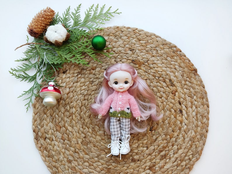 May include: A pink and green knitted sweater with a white collar and a green leaf design. The sweater is worn by a doll with pink hair and blue eyes. The doll is sitting on a brown woven mat.