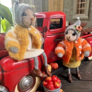 May include: Two plush toy animals wearing knitted hats and sweaters are sitting next to a red toy truck. The truck is filled with miniature pumpkins and apples. The animals are wearing a yellow and an orange sweater.