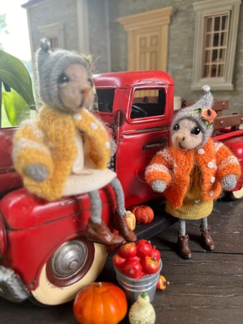 May include: Two plush toy animals wearing knitted hats and sweaters are sitting next to a red toy truck. The truck is filled with miniature pumpkins and apples. The animals are wearing a yellow and an orange sweater.