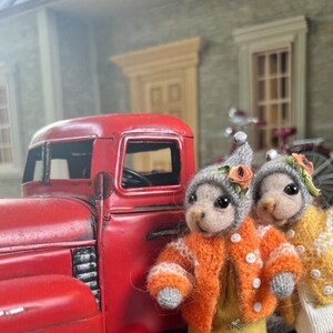 May include: Two small, gray, knitted animal figurines wearing orange and yellow sweaters with flower accents. The figurines are standing in front of a red toy truck.