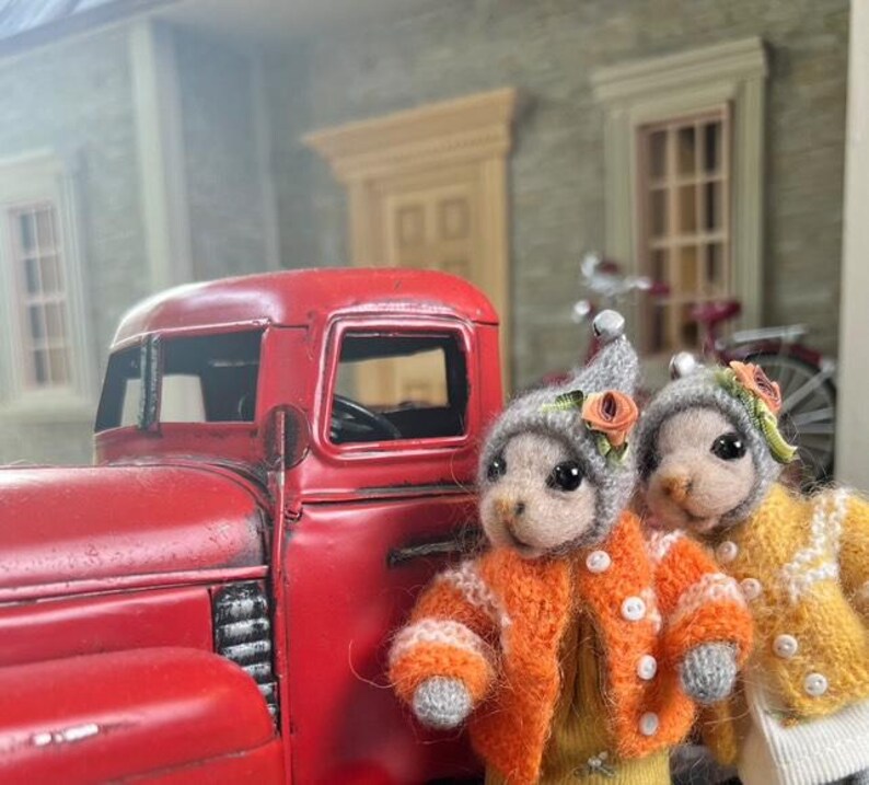 May include: Two small, gray, knitted animal figurines wearing orange and yellow sweaters with flower accents. The figurines are standing in front of a red toy truck.