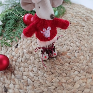 May include: A small, plush mouse wearing a red sweater with a white reindeer design and brown reindeer boots. The mouse is sitting on a woven rug.