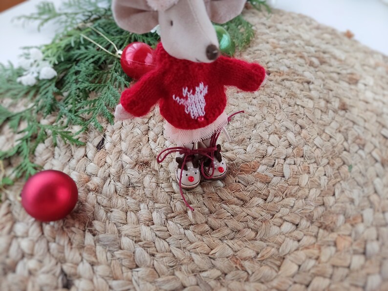 May include: A small, plush mouse wearing a red sweater with a white reindeer design and brown reindeer boots. The mouse is sitting on a woven rug.