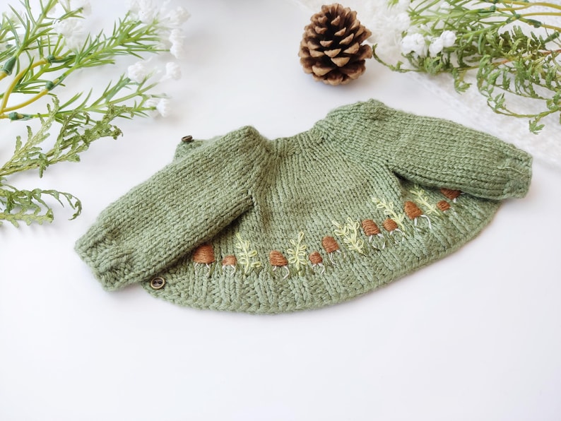 Op de afbeelding: Een saliegroene gebreide baby trui met lange mouwen. De trui heeft geborduurde bruine paddenstoelen en gele bladeren langs de onderste zoom. Twee kleine knopen zijn zichtbaar op de linkerschouder. Een dennenappel en groen zijn op de achtergrond.