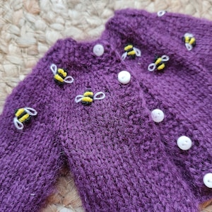 May include: A purple knitted cardigan with white buttons and embroidered bee accents. The bees are yellow and black with white wings. The cardigan is made of a soft, textured yarn.