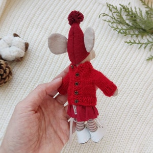Op de afbeelding: Een rode gebreide trui met een rode muts met pompon en witte schoenen. De trui heeft een knoopsluiting en de muts heeft een witte pompon bovenop.