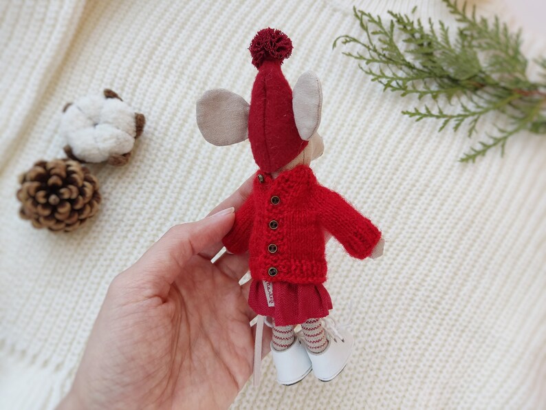 Op de afbeelding: Een rode gebreide trui met een rode muts met pompon en witte schoenen. De trui heeft een knoopsluiting en de muts heeft een witte pompon bovenop.