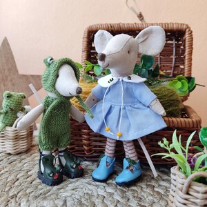 May include: Two handmade plush mouse toys, one wearing a green frog costume and the other wearing a blue dress with a white collar. Both mice are wearing blue shoes with red accents. The mice are sitting on a woven straw mat.