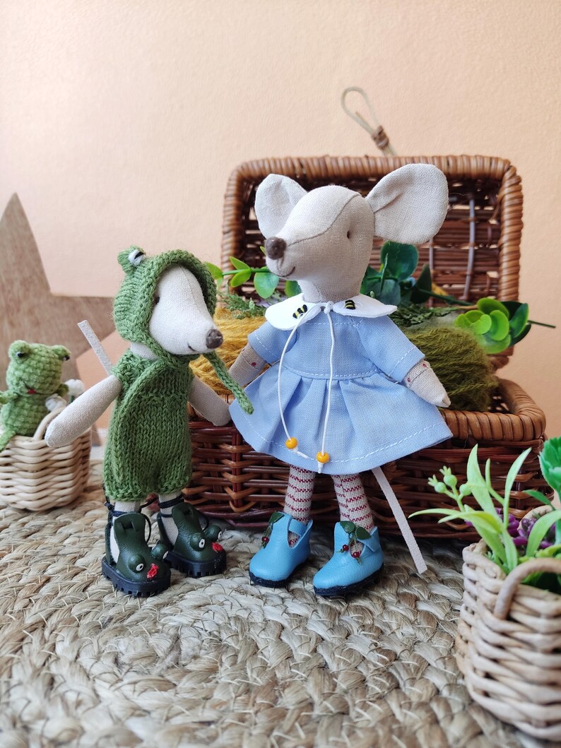 May include: Two handmade plush mouse toys, one wearing a green frog costume and the other wearing a blue dress with a white collar. Both mice are wearing blue shoes with red accents. The mice are sitting on a woven straw mat.