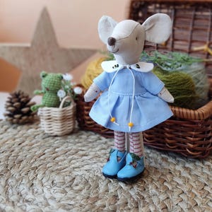 May include: A handmade plush mouse doll wearing a light blue dress with a white collar and red and white striped stockings. The doll is standing on a woven rug with a wicker basket and a green frog doll in the background.