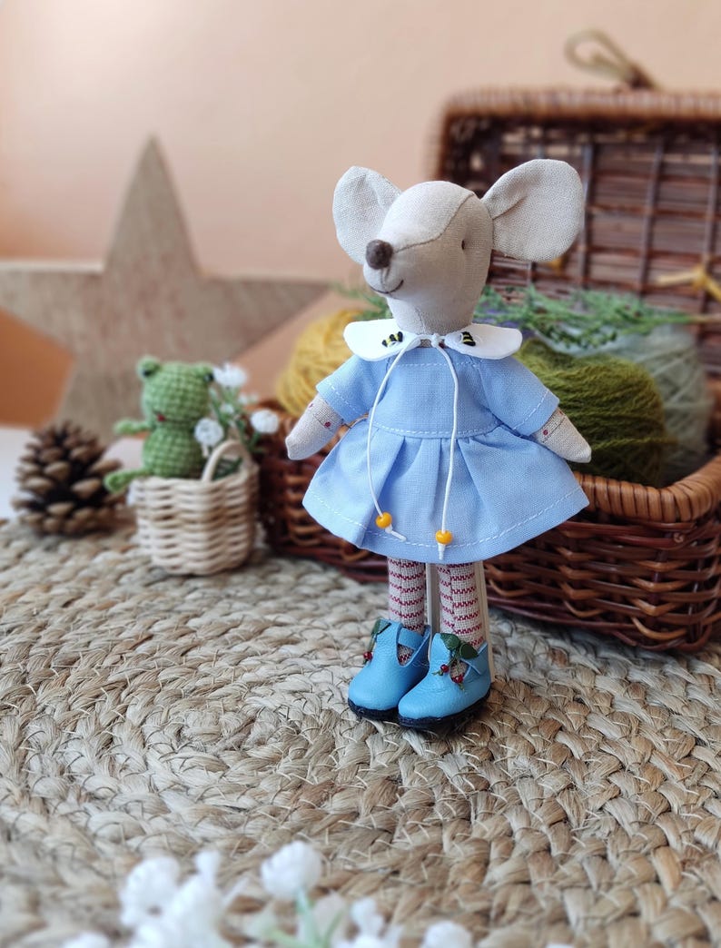 May include: A handmade plush mouse doll wearing a light blue dress with a white collar and red and white striped stockings. The doll is standing on a woven rug with a wicker basket and a green frog doll in the background.
