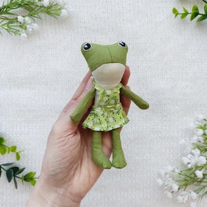 Könnte beinhalten: Eine handgefertigte grüne Froschpuppe mit einem hellgrünen und weißen Blumenkleid. Die Puppe hat große schwarze Augen und ist aus weichem Stoff gefertigt. Der Frosch wird in der Hand einer Person gehalten.