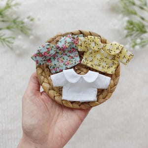 Puede incluir: Tres camisas de muñeca en miniatura en una cesta tejida. Una camisa es blanca con cuello Peter Pan, otra es azul claro con un estampado floral y la tercera es amarilla con un pequeño estampado floral blanco.