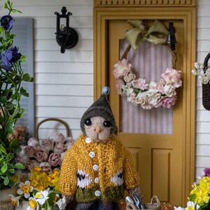 May include: A miniature doll wearing a yellow, green, and white knitted sweater and a gray hat stands in front of a dollhouse. The doll is holding gardening tools and is standing next to two wicker baskets filled with flowers.