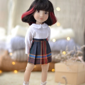 May include: A doll with black hair with red streaks, wearing a white collared shirt and a plaid skirt. The doll is wearing pink shoes and is standing on a wooden platform.